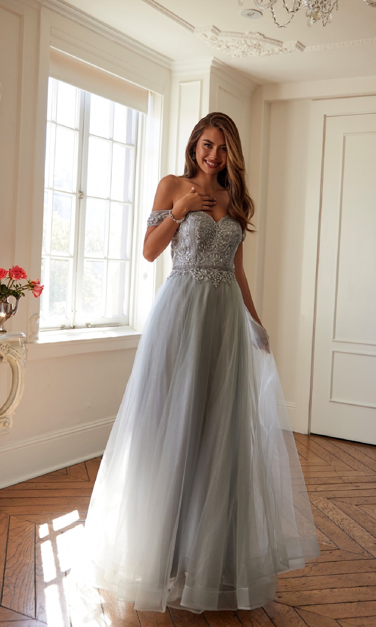 A woman wearing the Long Sweetheart Prom Ball Gown: Juliet 287, featuring a light gray tulle skirt and beaded corset bodice, stands in a bright room with wooden floors and a large window.